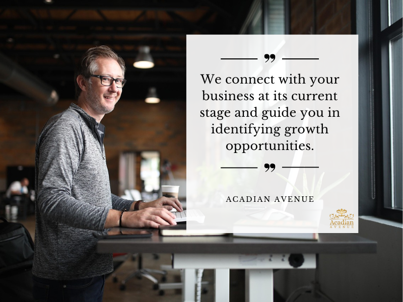 A person working at a standing desk with a laptop and coffee cup, set against a modern office background with brick walls and large windows. An overlay text reads, "We connect with your business at its current stage and guide you in identifying growth opportunities," attributed to Acadian Avenue with a logo in the bottom right corner. About Acadian Avenue